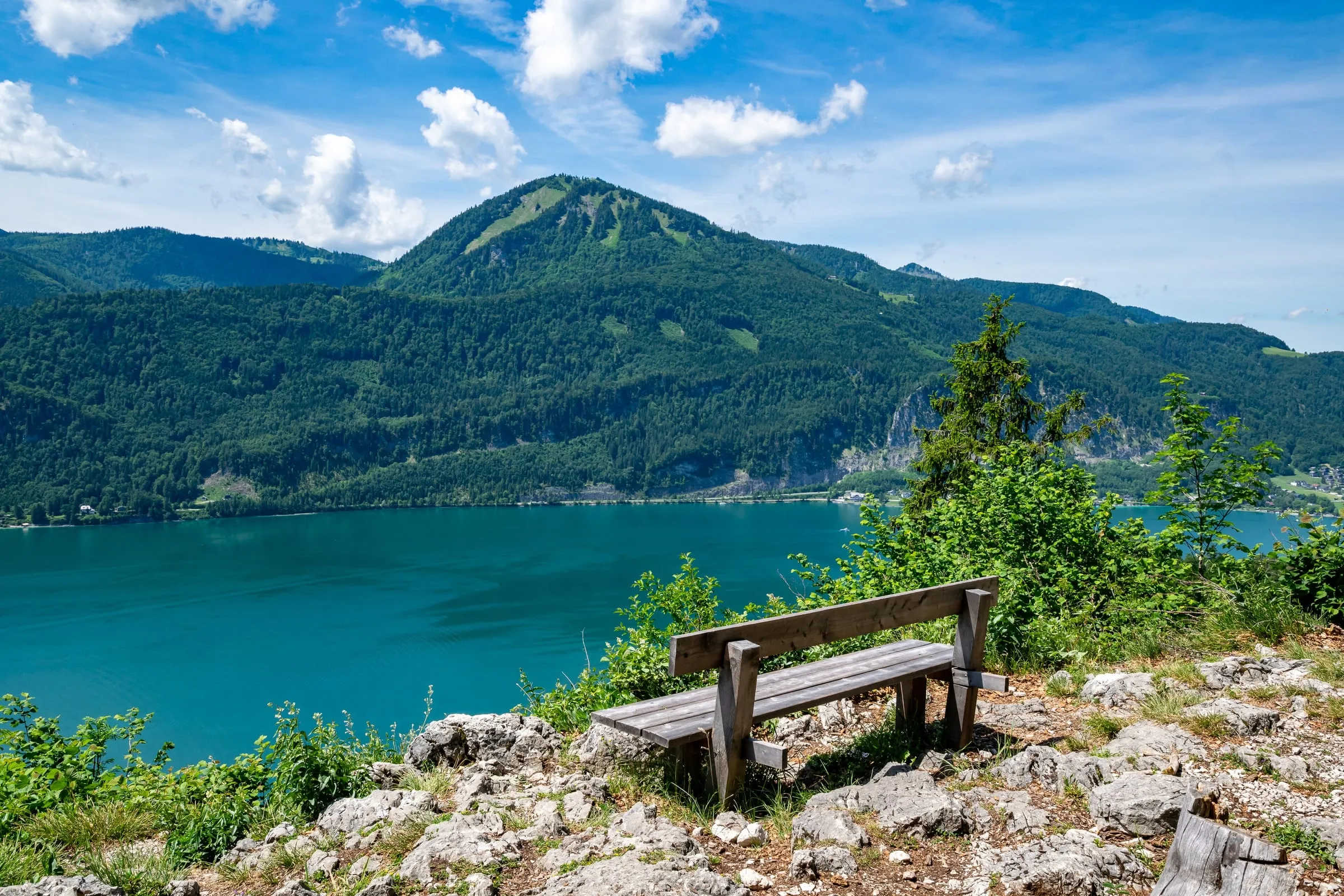 Eine Holzbank auf einem Felsen mit Blick über den türkisen Wolfgangsee und bewaldete Berge.