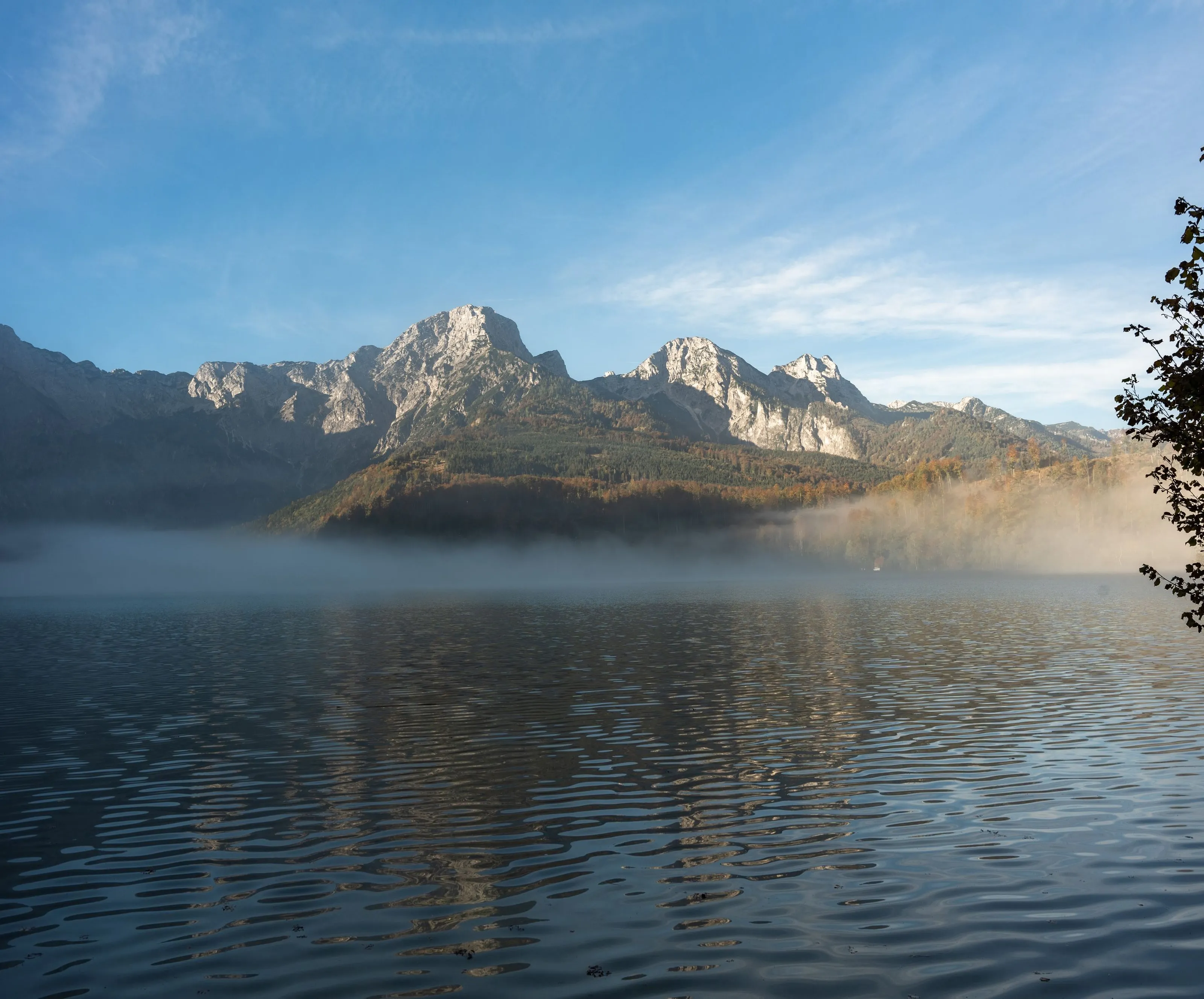 Morgendunst liegt über einem stillen Alpensee in Österreich, dahinter Felsgipfel im ersten Sonnenlicht.